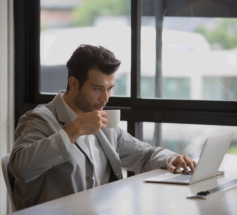 Man drinks cup of coffee while sitting behind a laptop in the studio of AllMediaLab web design.