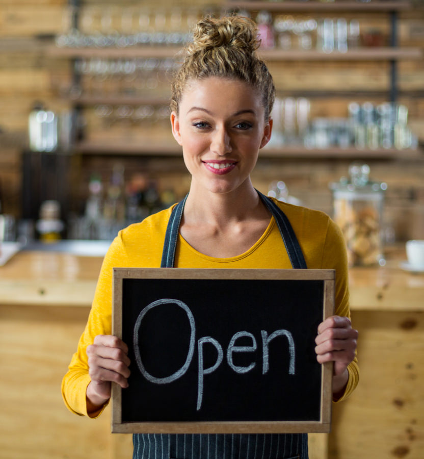 Female SME with blond hair stands in front of her catering company with a welcome sign in her hands